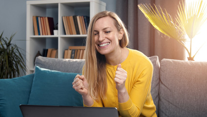 Overjoyed mature blond woman looking at laptop screen celebrating success sitting on sofa at home