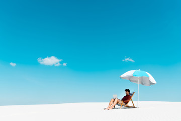 Obraz na płótnie Canvas freelancer sitting with laptop in deck chair under umbrella on sandy beach against blue sky