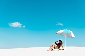 freelancer sitting with laptop in deck chair under umbrella on sandy beach against blue sky