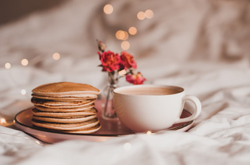 Tasty pancakes with dry roses and cup of coffee in bed over glowing lights. Good morning.