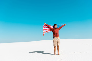 happy man with american flag on windy sandy beach against clear blue sky