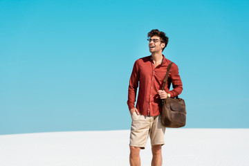 smiling handsome man with leather bag on sandy beach against clear blue sky