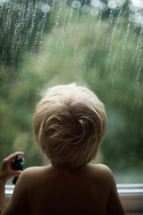 The boy looks out the window where raindrops are falling.