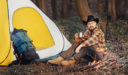 Hiker drinking hot tea sitting next to campfire and yellow and white tent enjoying spring morning