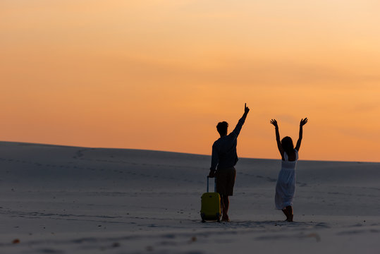 back view of couple walking on beach with hands in air and travel bag at sunset