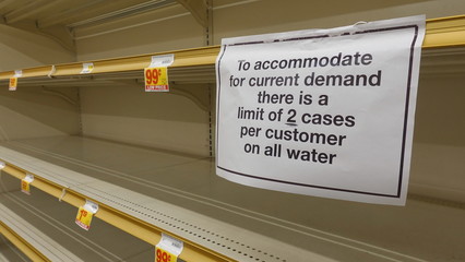 Bare water shelves in a grocery store, and a sign limiting purchases. People are hoarding food to...