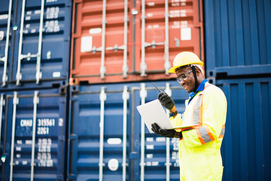 Adult African American Men Worker Check And Control Loading Freight Containers By Use Computer Laptop At Commercial Shipping Dock Smiling Felling Happy. Cargo Freight Ship For Import Export Concept