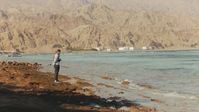 Pretty woman walking alone on stony shore beach near sea, the waves are breaking on the shore, Egypt Sinai mountain on the background, slow motion, 4k