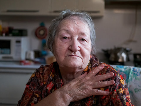 Funny old woman with gray hair and wrinkled face. pensioner suffers from senile memory loss and is upset because of her dependence on relatives and carers. Woman sitting at table in kitchen