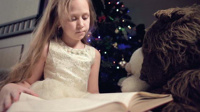 A little blonde girl in a festive dress with a book in her hands sits next to soft toys against the background of a Christmas tree and reads a book leading the page with her place.