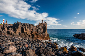 Young man standing on a rock bluff looking into the blue ocean. At background lighthouse and clouds.