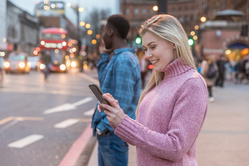 Smiling woman in London using phone near a busy road - blonde woman and black man on background in the city at dusk waiting for a bus or taxi