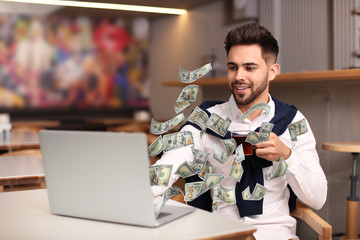 Man with modern laptop and flying dollar banknotes at table indoors. People make money online