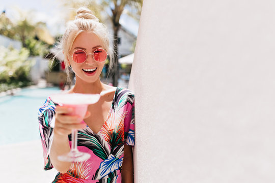 Beautiful woman in stylish pink glasses tasting fruit beverage. Outdoor photo of emotional caucasian girl in trendy clothes laughing and drinking cocktail.