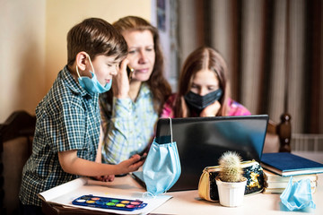 A woman and her children study through a laptop online because schools are closed due to quarantine. Focus on medical mask hanging on computer. The concept of distance education