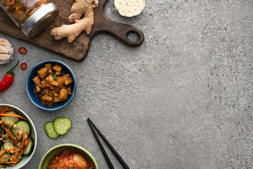 top view of tasty kimchi in bowls and jar near cutting board, chopsticks, garlic, ginger and chili pepper on concrete surface