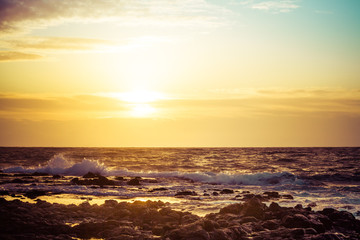 The sun rising over the ocean with a rocky foreground and rough surface crashing into the shore of Hawaii.