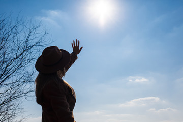 Silhouette of a girl in a hat and coat on a background of the sky with clouds and the sun.