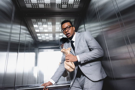 Nervous African American businessman with paper bag suffering from panic attack in elevator