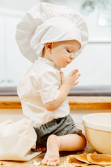 Side view of a naughty little boy in white cook suit sitting on the kitchen table barefoot and kneading dough for fritters. Concept of natural curiosity of children