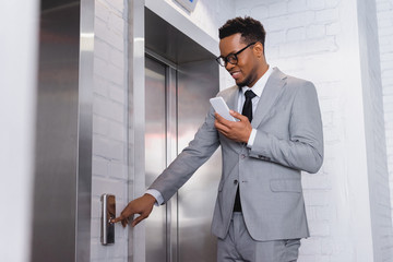 positive african american businessman using smartphone while pushing elevator button