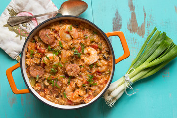 Jambalaya in an orange cast iron pot. Top view on rustic turquoise table, shown with bunch of scallions. 