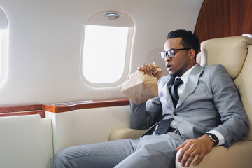 Worried african american businessman breathing with paper bag while having panic attack during flight on private plane