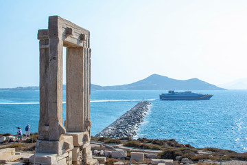 Image shows the famous Portara monument at Naxos island with people taking photos of it.A ship is cruising at the blue sea at the background.