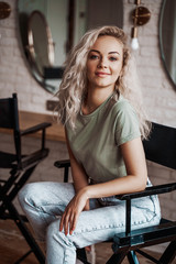 Obraz na płótnie Canvas Cool and smiling young female model with blond curly hair posing on a make up chair in a bright room in front of mirrors, looking cute and interested