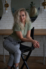 Obraz na płótnie Canvas Cool and smiling young female model with blond curly hair and white boots posing on a make up chair in a bright room in front of mirrors, looking cute and interested