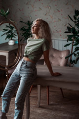 Attractive and cheerful young woman wearing bright clothes and high heels posing in a bright apartment leaning on wooden table