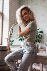 Fototapeta na wymiar Attractive young woman with blonde curly hair wearing casual clothes posing in a bright apartment leaning on wooden table and holding her hand up