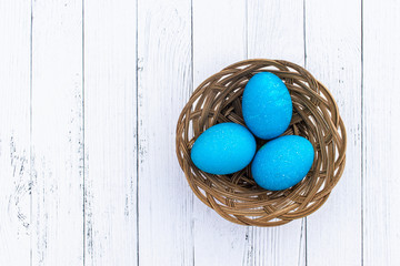Three small blue Easter eggs in a small basket on light white wooden background with copy space