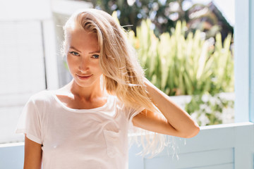 Playful blonde girl posing near window. Beautiful european lady in trendy white t-shirt relaxing in sunny morning