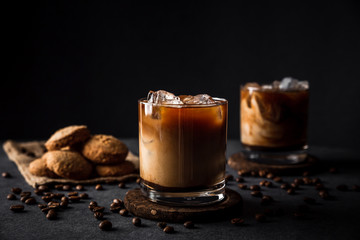 A horizontal photo of 2 rocks glasses with cold iced coffee with milk, coffee beans around, cookies, dark background, deep shadows - 334566946