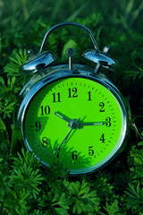 Green clock on green grass background. Vintage alarm Clock outdoors. Abstract seasonal background.
