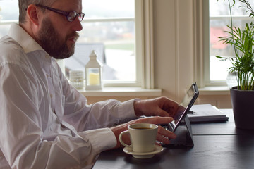 Freelancer bearded man 50 years old in a white shirt sits at a table and takes notes in a laptop. Concept: freelancer at home behind a laptop screen, remote work in quarantine.