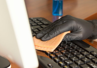 hand in a black nitrile glove wipes the keyboard on the workplace with an orange napkin