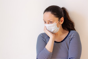 A girl in a striped sweater and medical mask thoughtfully props her head with her hand and looks toward the copy space. European appearance. Long black hair. White background. Horizontal.