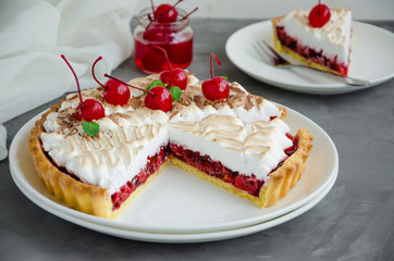 Tart with cherry filling and Italian meringue with a cocktail cherry, chocolate and mint on top on a white plate on a dark concrete background. Cherry pie. Horizontal orientation, close up.