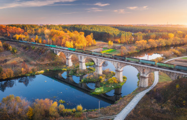 Aerial view of moving freight train on railroad bridge and river at sunset in autumn. Landscape with bridge, wagons, water, colorful trees, sky with gold sunlight in fall. Top view of railway station