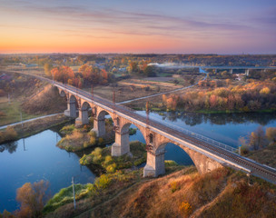 Obraz premium Aerial view of railroad bridge and river at beautiful sunset in autumn. Industrial landscape with bridge, blue water, colorful trees, rural railway station, orange sky in fall at dusk. Top view