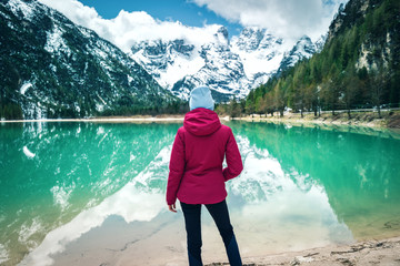Young woman in red jacket is standing on shore of lake with azure water at sunny day in spring. Landscape with girl, reflection in water, snowy mountains, blue sky with clouds, green trees. Travel