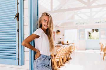 Attractive tanned lady with gorgeous smile posing in cafeteria early in morning. Photo of cute young woman in vintage outfit standing in restaurant