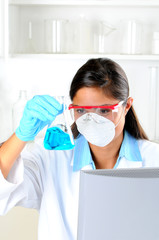 Naklejka premium Young female Lab Tech seated behind computer monitor holding up a flask of blue chemicals in laboratory setting. Vertical format.