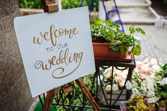 there is a sign that says "Welcome to our wedding", on the easel  on a background of greenery and flowers