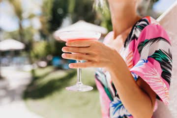 Photo of tanned female hand holding glass of fruit cold beverage. Stylish young woman drinking cocktail while resting outdoor.