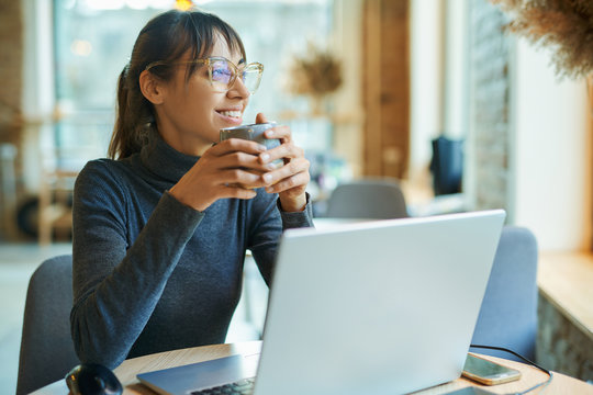 Attractive smiling woman enjoying coffee while working in cafe or coworking space. sitting at the desk with laptop looking at window. Concept remote work, freelance, using laptop computer or net-book.