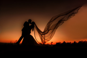 Silhouette of bride and groom kissing at sunset. The veil is flying.