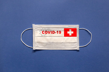 Flag of Switzerland and inscription COVID-19 on a medical mask on a blue background. Healthcare and medical concept. Pandemic virus COVID-19